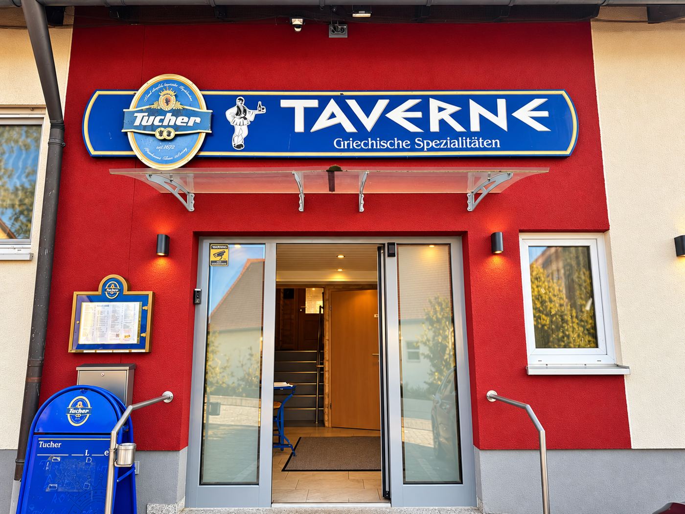 Eingang der Taverne Sonnenblick mit rotem Giebel und Schild „Taverne – Griechische Spezialitäten“